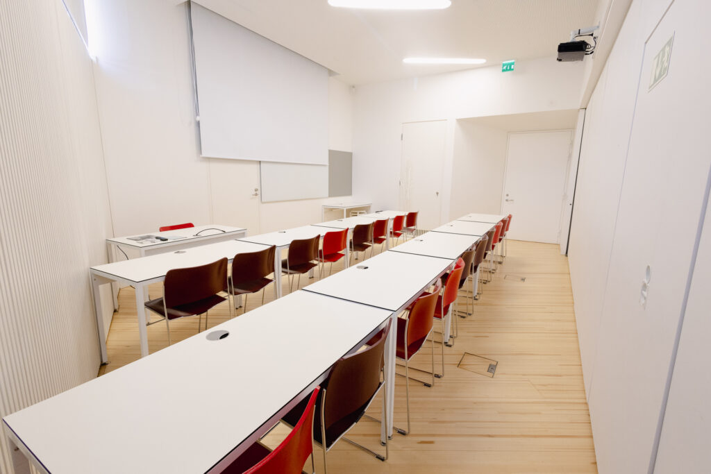 Pieni sali, jossa valkokangas sekä kaksi riviä valkoisia pöytiä ja punaisia tuoleja