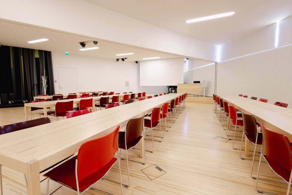 Iso sali, jossa 4 riviä pöytiä ja tuoleja. Taustalla valkokangas.